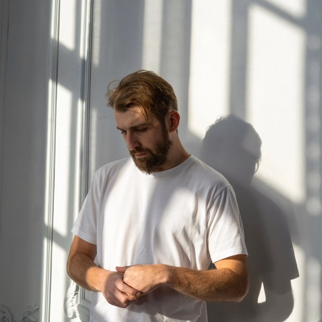 Man standing quietly by a sunlit window, looking down